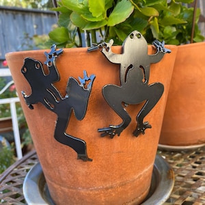 May include: Two black metal frog silhouettes designed to hook onto the rim of a terracotta plant pot. Each frog figure features a flat, cutout design with detailed limbs and toes, positioned to appear as if they are climbing the side of the pot.