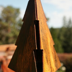 May include: A hanging metal ornament shaped like a stylized evergreen tree with a weathered, rusted brown finish. The structure is composed of flat, angular panels with visible vertical gaps and a metal ring at the top for suspension purposes.