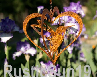 May include: A rusty metal hummingbird wind chime with a heart-shaped frame. The hummingbird is inside the heart, and the wind chime hangs from a chain. The background features purple flowers and green foliage. The text "RusticSun19" is visible.
