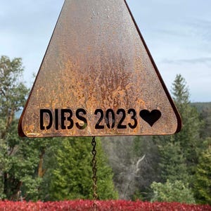 May include: A rusty metal triangle wind chime with the text "DIBS 2023" and a heart. The chime has a rusty metal heart hanging from it.
