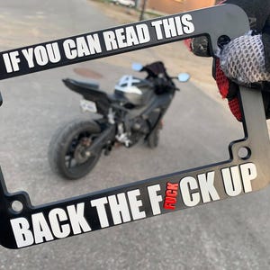 May include: Black license plate frame with white text that reads "IF YOU CAN READ THIS" at the top and "BACK THE FUCK UP" at the bottom. The word "FUCK" is in red. A motorcycle is visible in the background.