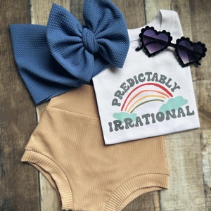 May include: A baby outfit set featuring a blue bow headband, a white t-shirt with a rainbow graphic and the text "Predictably Irrational", tan shorts, and heart-shaped sunglasses.