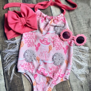 May include: Pink baby onesie with a white fringe trim and a white and pink cactus print. The onesie has the text "What would Dolly do?" printed on it. The onesie is paired with a pink headband and pink sunglasses.