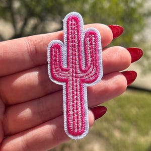 May include: A pink cactus patch with white trim is held up against a blurred green background. The patch is a stylized representation of a cactus plant, with a detailed embroidered design.