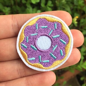 Könnte beinhalten: Gestickter Aufnäher in Form eines lila Donuts mit blauen Streuseln und einem weißen Zentrum. Der Donut hat eine goldfarbene Umrandung. Der Aufnäher befindet sich vor einem Hintergrund aus grünem Laub.