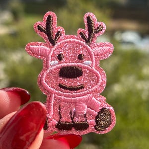 Pode incluir: Um patch bordado rosa em forma de rena. A rena tem chifres castanhos, um nariz castanho e um rosto sorridente. O patch é segurado por uma mão com verniz de unhas vermelho.