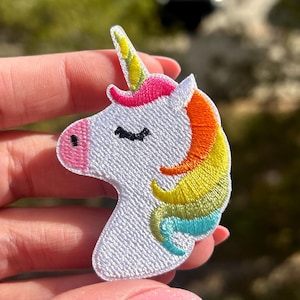 May include: A white unicorn patch with a pink nose, a yellow horn, and a rainbow mane in shades of pink, orange, yellow, and blue. The patch is held in a hand against a blurred outdoor background.