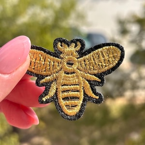 May include: Embroidered bee patch with gold thread detailing on a black background. The bee has outstretched wings and a detailed body. This patch can be sewn or ironed onto clothing or accessories.