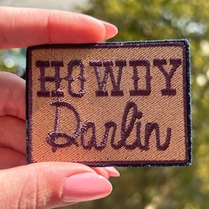 May include: A rectangular patch with the words "HOWDY Darlin" in a dark brown, cursive font. The patch has a tan background and a dark brown border. The patch is held by a hand with pink nail polish.