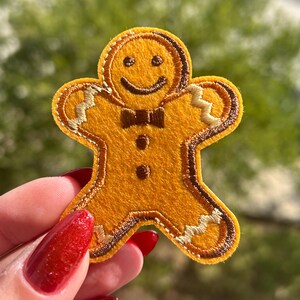 May include: A gingerbread man patch with a smiling face, bow tie, and button details. The patch is a warm yellow color with brown and tan accents, perfect for holiday crafts and apparel.