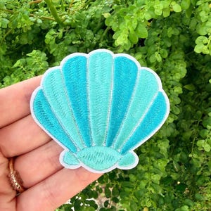 Puede incluir: Un parche de concha marina turquesa y verde azulado con ribete blanco. El parche bordado presenta un diseño detallado de una concha marina con líneas radiales. El parche se sostiene frente a un fondo de follaje verde.