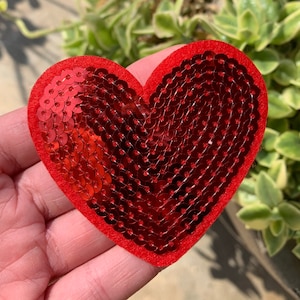 May include: A red heart-shaped patch with a sequined surface. The patch is held in a hand, with a green leafy background. The heart is approximately 3 inches wide and 3 inches tall.