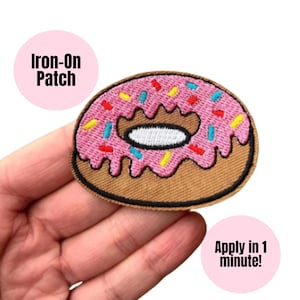 Op de afbeelding: Geborduurde donut patch met roze glazuur en kleurrijke hagelslag. De patch heeft een bruine rand en de tekst "Iron-On Patch". De patch is ontworpen om in 1 minuut te worden aangebracht.