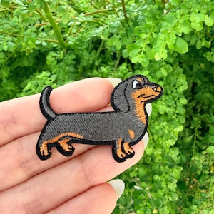 May include: Embroidered patch of a dachshund dog in black, orange, and white. The dog is in a side profile, with its body and legs outlined in orange. The patch is held against a background of green foliage.