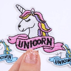 May include: Embroidered unicorn patch with a white body, a spiraled horn, and a pink, purple, and yellow mane. The patch has a pink banner with the word "UNICORN" in black letters. The patch is on a white background.
