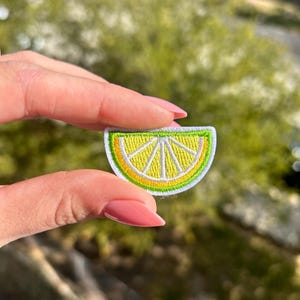 May include: Embroidered patch in the shape of a lime slice. The patch features a yellow center with white stitching, a green rind, and a white border. The patch is held up against a blurred green background.