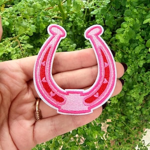 May include: A pink horseshoe patch with a white border. The horseshoe is filled with red hearts and has a light pink center. The patch is held in front of a background of green foliage.