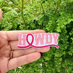 Puede incluir: Un parche bordado con la palabra HOWDY en letras de bloque de color rosa claro. La letra O está reemplazada por un lazo de concienciación de color rosa brillante que se extiende en una línea curva bajo el texto, sobre fondo blanco.
