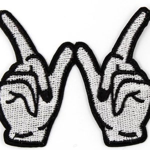 May include: Embroidered patch showing a white hand gesture with the index and middle fingers raised, forming a 'V' shape. The patch has a black outline and is on a white background. The design is a symbol of peace or victory.