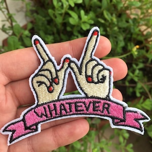 May include: Embroidered patch featuring two hands forming the 'W' symbol, with red nail polish. A pink banner below reads 'WHATEVER' in black letters. The patch has a white border and is set against a green leafy background.