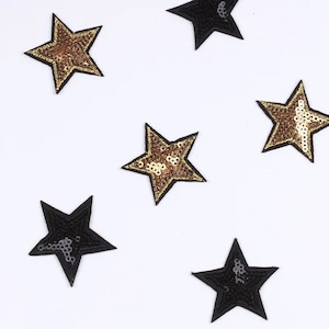 Puede incluir: Un conjunto de parches en forma de estrella. Algunas estrellas son negras con lentejuelas plateadas, mientras que otras son doradas con lentejuelas doradas. Cada estrella tiene un borde negro. Los parches están dispersos sobre un fondo blanco.