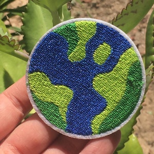 May include: A round embroidered patch featuring a stylized Earth design. The patch has a blue ocean and green continents, with a white border. The patch is likely for clothing or accessories.