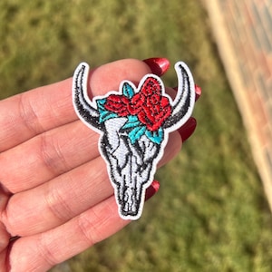 May include: Embroidered patch featuring a white bull skull with black horns. The skull is adorned with red roses and turquoise leaves. The patch has a white border and is likely for clothing or accessories.