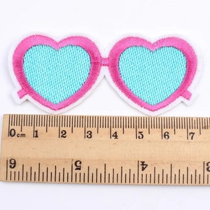 May include: Embroidered patch of heart-shaped sunglasses. The sunglasses have pink frames, light blue lenses, and a white border. The patch is approximately 3 inches wide.