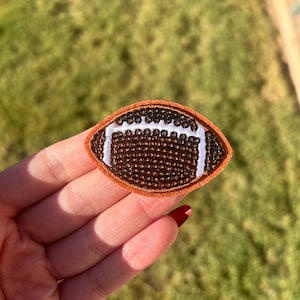 May include: An embroidered patch in the shape of a football. The patch has a brown background with a textured pattern, white stripes, and a brown border. The patch is held in a hand against a blurred green background.