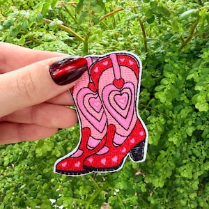 May include: Embroidered patch featuring a pair of pink and red cowboy boots with heart designs. The boots have black heels and are set against a backdrop of green foliage. The patch is a decorative item.
