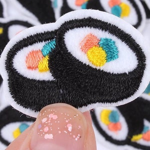 May include: Embroidered sushi roll patch with black nori, white rice, and colorful fillings of orange, yellow, and teal. The patch has a white border and is likely for decorative purposes, such as on clothing or accessories.