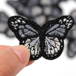 Op de afbeelding: Geborduurde vlinderpatch in zwart en grijs. De vlinder heeft gedetailleerde vleugelpatronen met witte en grijze accenten. De patch wordt in een hand gehouden, met andere patches wazig op de achtergrond.