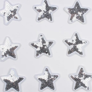 May include: Nine star-shaped patches with silver sequins and white borders are arranged on a white background. The sequins reflect light, creating a shimmering effect. These patches are suitable for various craft projects, such as embellishing clothing or accessories.