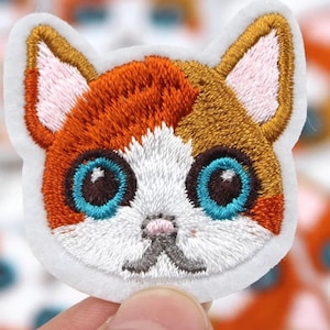 May include: Embroidered cat face patch. The cat has orange, brown, and white fur, with large blue eyes and a pink nose. The patch is outlined in white.