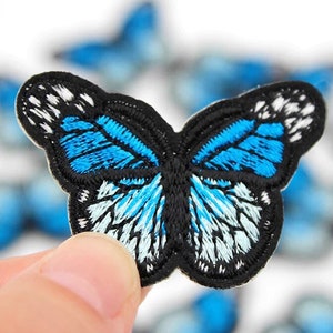 May include: Embroidered butterfly patch with a black outline and vibrant blue and white wing details. The butterfly patch is a decorative item, perfect for adding to clothing or accessories. The patch is held in a person's hand.