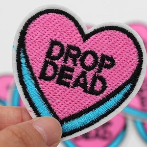 May include: Embroidered heart-shaped patch with the words "DROP DEAD" in black on a pink background. The patch has a white border with a blue accent. The patch is held by a hand.