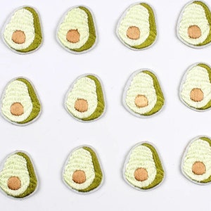 May include: A collection of twelve embroidered avocado patches. Each patch features a green rind, a creamy white interior, and a brown seed. The patches are arranged in a grid pattern against a white background.