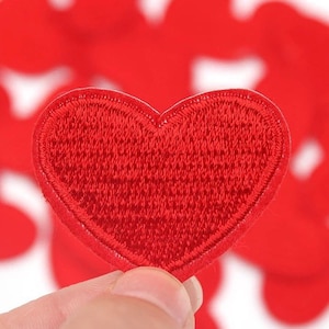 May include: A close-up of a red heart-shaped embroidered patch. The patch is made of red thread and has a detailed, textured surface. The heart is held in a person's hand, with other similar patches blurred in the background.