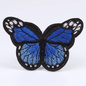 May include: Embroidered butterfly patch featuring a detailed design. The butterfly has blue wings with black outlines and white accents. The patch is suitable for clothing, bags, or other fabric items.