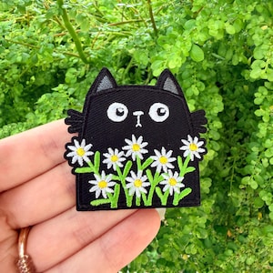 May include: Black embroidered patch of a cartoon cat with white eyes and ears, and small wings. The cat is surrounded by white daisies with yellow centres and green stems. The patch is held in front of a background of green foliage.