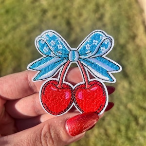 May include: Embroidered patch featuring two red cherries with a blue bow. The cherries have a glossy finish and are connected by stems. The bow is light blue with white lace detailing. The patch is on a white background.