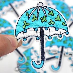 Puede incluir: Parche de paraguas bordado con dosel azul claro, ribete blanco y mango negro. El dosel presenta diseños verdes en forma de trébol. El parche se sostiene sobre un fondo de parches similares.