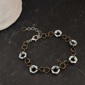May include: A unique bracelet featuring silver-colored hex nuts linked together with a bronze-toned chain. The bracelet has a lobster clasp closure and an adjustable chain. The industrial-inspired jewelry is set against a dark background.