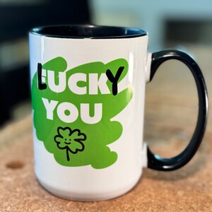 May include: White ceramic mug with a black handle and black rim. The mug has a green and white design with the text "LUCKY YOU" and a green four-leaf clover with a smiley face.