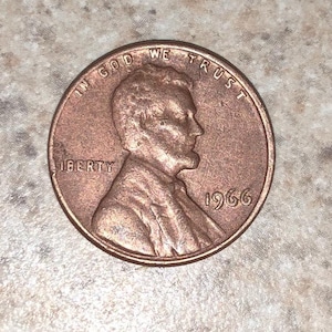 Puede incluir: Un centavo estadounidense de cobre de 1966 con las palabras "In God We Trust" y "Liberty" alrededor del borde. El año 1966 está impreso en la parte inferior derecha de la moneda.