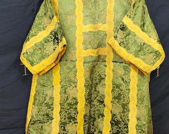 May include: Green and gold brocade garment with floral patterns. The garment has long sleeves and is trimmed with gold borders. It is hanging on a green hanger against a dark background. The fabric has a sheen.