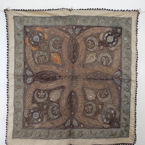 May include: A square textile with an intricate, symmetrical design in shades of brown, beige, green, and orange. The central pattern features floral and abstract shapes. The border is a lighter beige with a green trim, and the edges are finished with a dark beaded trim.