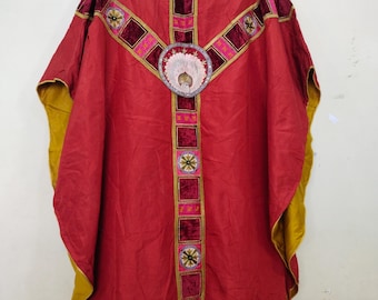 May include: A red liturgical vestment with gold and pink accents. The garment features a cross design with embroidered details and a central circular emblem. The edges are trimmed with gold and yellow fabric.
