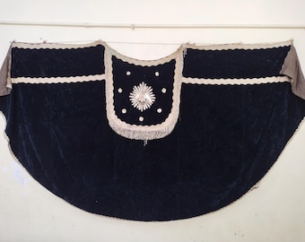 May include: A dark blue velvet cape with white trim and a decorative central panel. The panel features a floral design with white embroidery and fringe. The cape has a scalloped edge and is likely a theatrical or ceremonial garment.