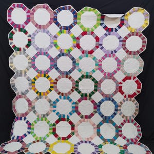 May include: A quilt featuring a repeating pattern of white octagonal fabric centers surrounded by borders made of various colorful patterned fabric strips. The white sections have a quilted swirl design, and the edges are finished with white binding.